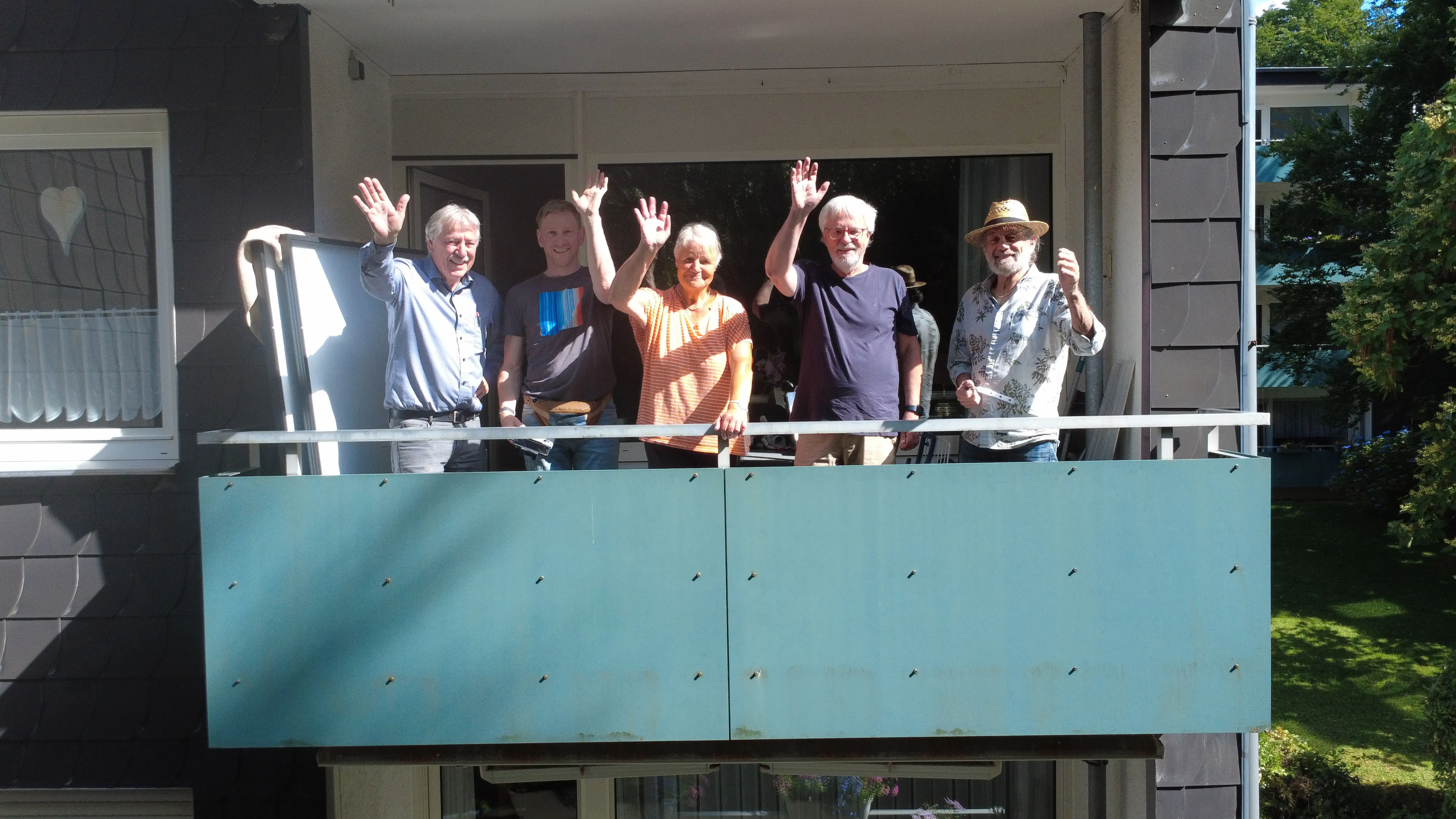Fünf Personen winken vom Balkon eines schiefergedeckten Wohnhauses; die Balkonbrüstung trägt noch die grüne Blende ohne Solarmodule