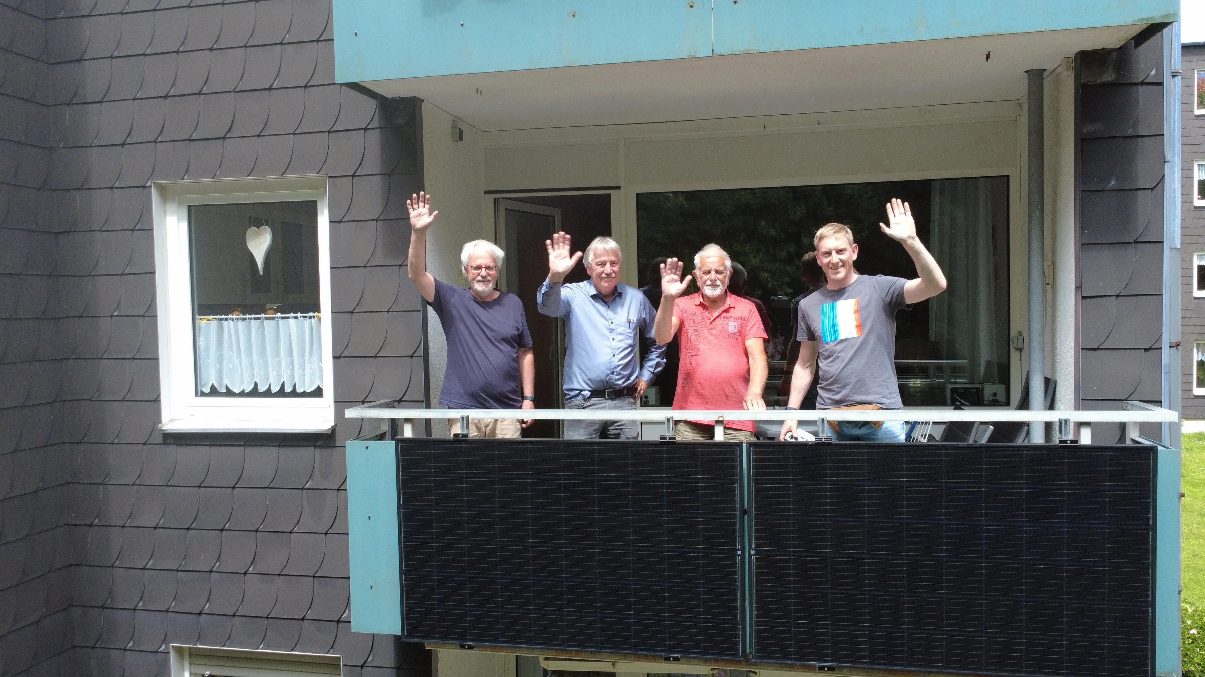 Vier Personen winken vom Balkon; an der Brüstung hängen zwei schwarze Solarmodule des Balkonkraftwerks