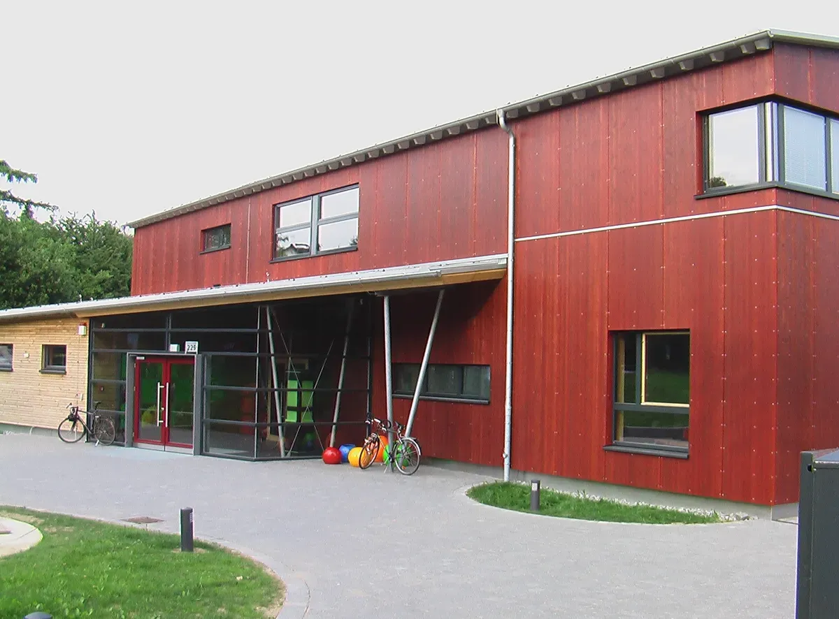 Zweigeschossiges Gebäude mit rot gestrichener Holzfassade und überdachtem Glaseingang, davor abgestellte Fahrräder