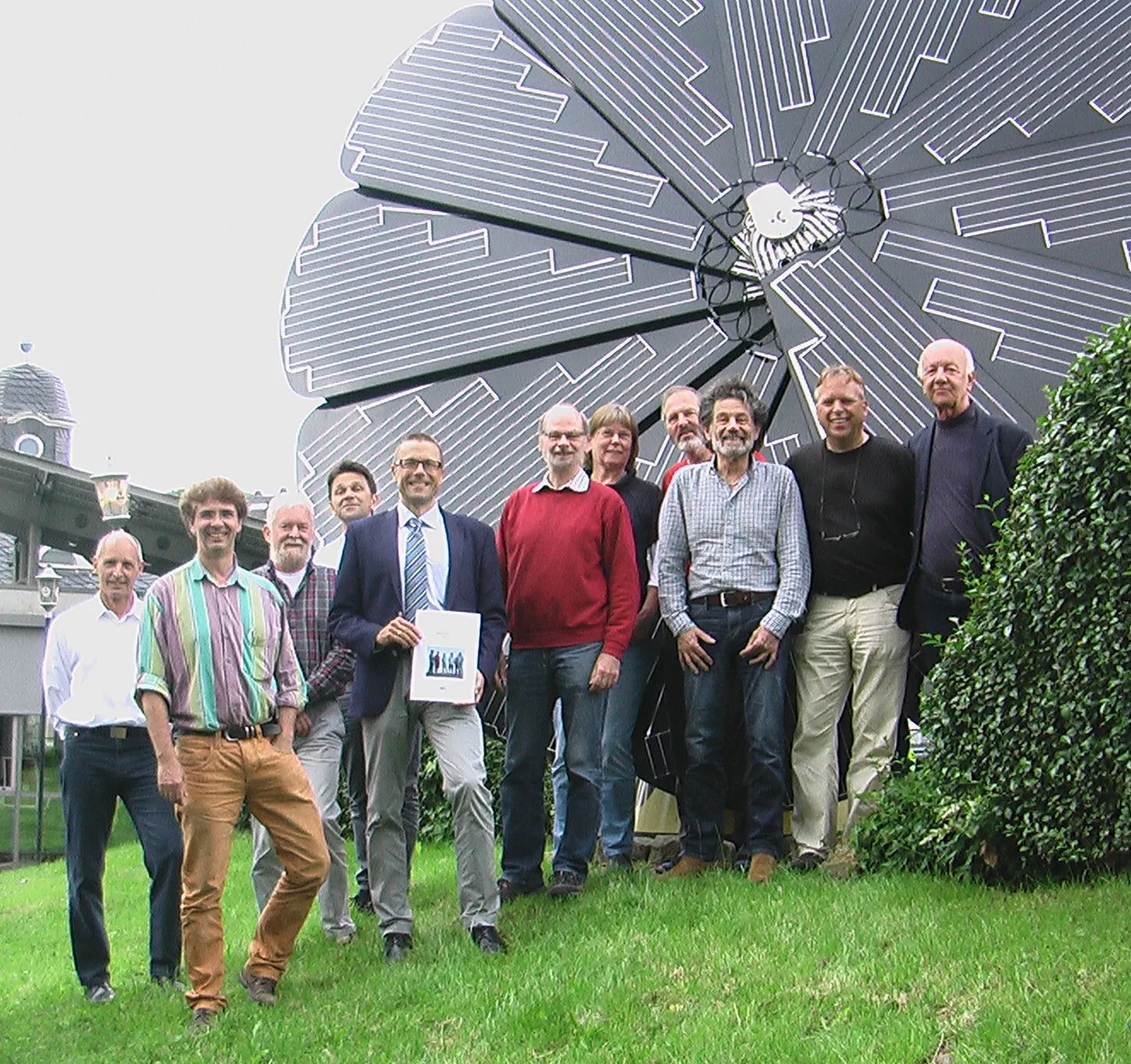 Elf Personen stehen nebeneinander auf einer Wiese vor einem großen blütenförmigen Solarmodul; ein Mann im Anzug in der Mitte hält eine Urkunde in den Händen.